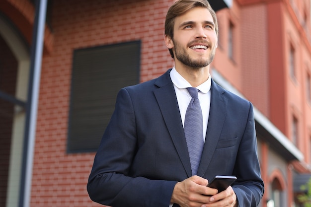 Retrato de un hombre de negocios joven sonriente vestido con traje hablando por móvil de pie al aire libre.