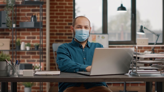 Foto retrato de un hombre de negocios con mascarilla y portátil en la oficina. empresario mirando la cámara y trabajando con la computadora para planificar el proyecto y la presentación durante la pandemia de covid 19