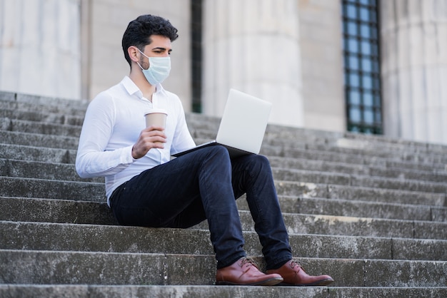 Retrato de hombre de negocios con mascarilla y usando su computadora portátil mientras está sentado en las escaleras al aire libre.