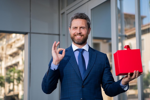 Retrato de hombre de negocios con ok firmar en traje mostrando caja de regalo