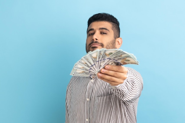 Retrato de un hombre de negocios rico y barbudo, confiado en sí mismo, sosteniendo billetes de dólar en la cámara, jactándose de dinero ganado en la lotería, usando una camisa a rayas, tiro de estudio interior aislado en fondo azul