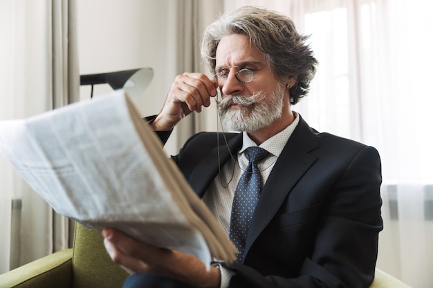 Retrato de un hombre de negocios senior canoso guapo concentrado con anteojos en el interior en casa vestido con ropa formal leyendo el periódico.