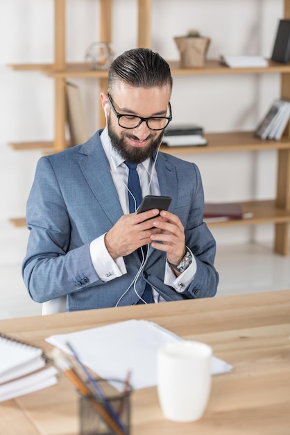 Retrato de un hombre de negocios sonriente escuchando música y usando un teléfono inteligente con auriculares en el lugar de trabajo