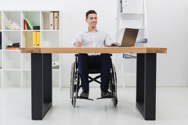 Foto retrato de un hombre de negocios sonriente que se sienta en la silla de ruedas usando la computadora portátil en el lugar de trabajo