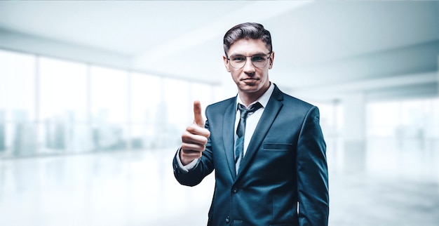 Retrato de un hombre de negocios con traje. Está de pie en la oficina de un rascacielos y muestra un pulgar hacia arriba. Concepto de negocio.