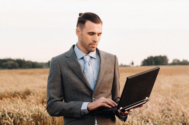 Retrato de un hombre de negocios en un traje sosteniendo una computadora portátil en un campo de trigo con el telón de fondo de un molino de viento y el cielo de la tarde