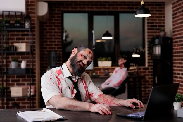 Retrato de un hombre de negocios zombi sentado en el escritorio, luciendo espeluznante y aterrador en la oficina de la empresa. Monstruo cruel que come cerebro con heridas sangrientas trabajando en una laptop, horrible diablo poseído.