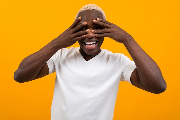 Retrato de un hombre negro africano guapo rubio cerrando los ojos con las manos en una camiseta blanca sobre un fondo amarillo studio