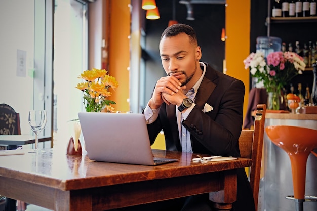 Retrato de hombre negro elegante usando una computadora portátil en un restaurante.