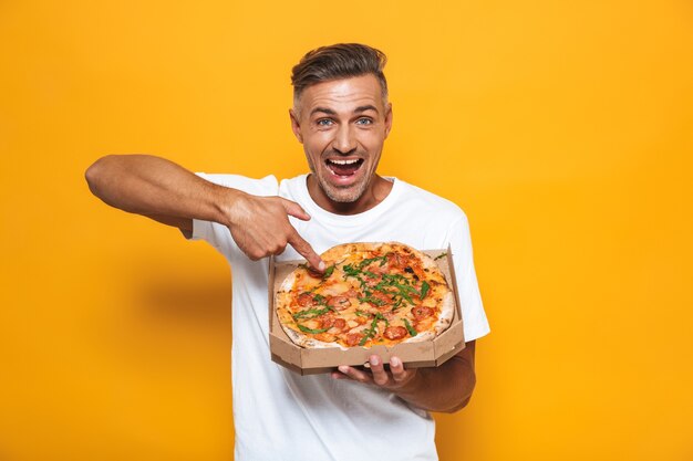 Retrato de hombre optimista de 30 años en camiseta blanca sosteniendo y comiendo pizza mientras está de pie aislado en amarillo