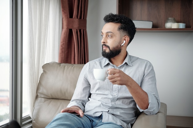 Retrato de hombre pensativo bebiendo una taza de café por la mañana en casa escuchando música y mirando a otro lado