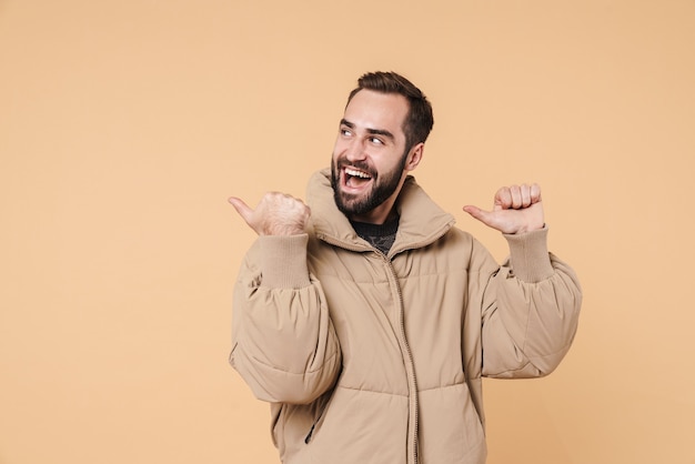 Retrato de hombre positivo en chaqueta de invierno sonriendo y señalando con el dedo a un lado en copyspace aislado en beige