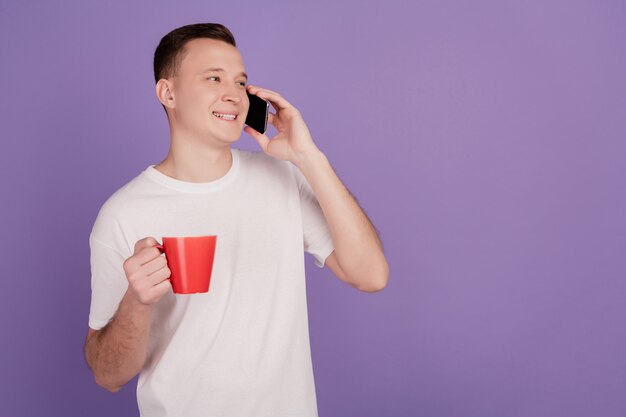 Retrato de hombre positivo mantenga conversación telefónica mirar espacio vacío beber té sobre fondo púrpura