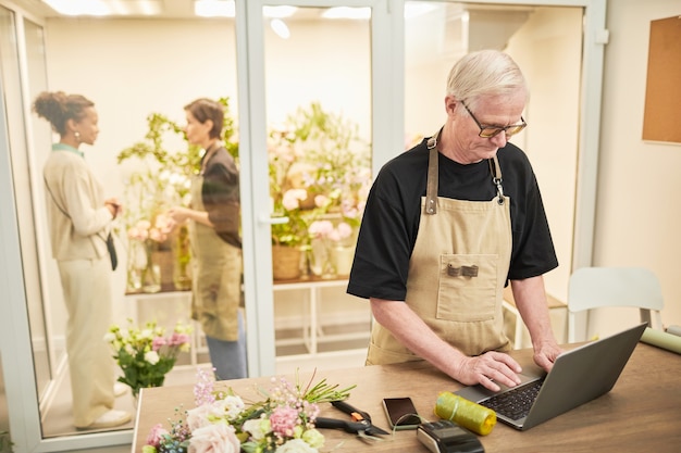 Retrato de hombre propietario de una pequeña empresa usando una computadora portátil y con una máscara mientras administra la copia de la tienda de flores ...