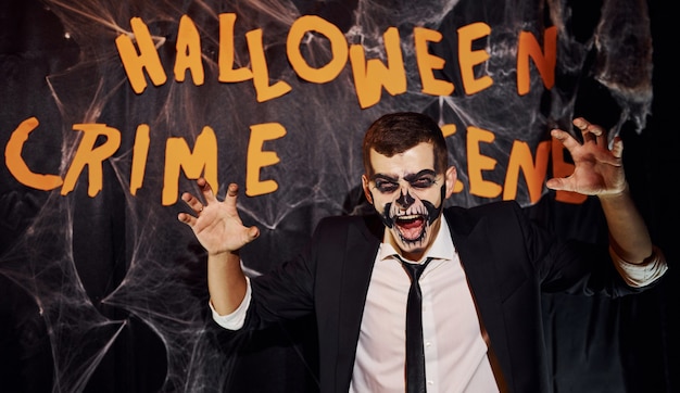 Retrato de hombre que está en la fiesta temática de halloween con maquillaje de esqueleto aterrador y disfraz contra la pared de la escena del crimen.