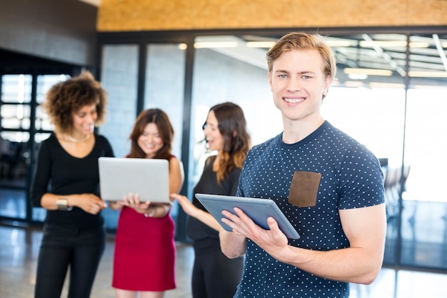 Retrato del hombre que sonríe y que sostiene la tableta digital mientras que sus colegas que se colocan detrás de él en oficina