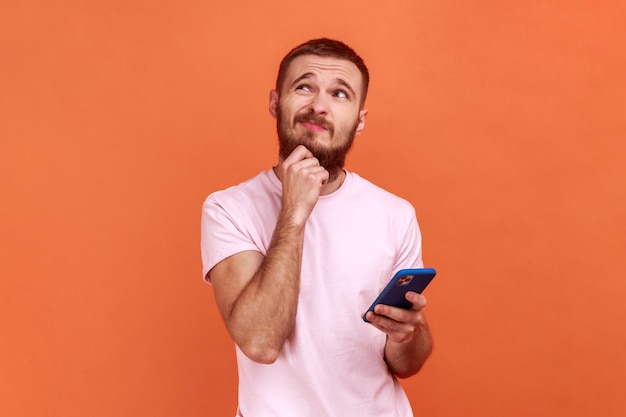 Retrato de un hombre que sostiene la barbilla de pie con un teléfono inteligente en la mano contemplando la actualización del software eligiendo tarifas adecuadas usando una camiseta rosa Foto de estudio interior aislada en un fondo naranja