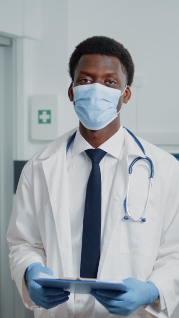 Retrato de un hombre que trabaja como médico parado en la sala del hospital, usando mascarilla contra la pandemia del coronavirus. Médico con estetoscopio y bata blanca con documentos de chequeo.