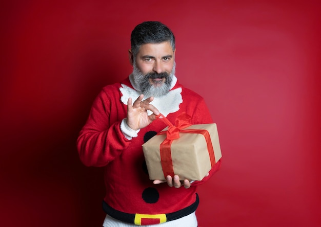 Retrato de un hombre que viste un suéter de Navidad abriendo una caja de regalo aislada en rojo