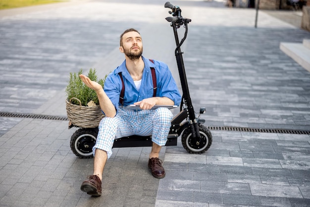 Retrato de un hombre raro y elegante sentado con una laptop en una scooter eléctrica al aire libre