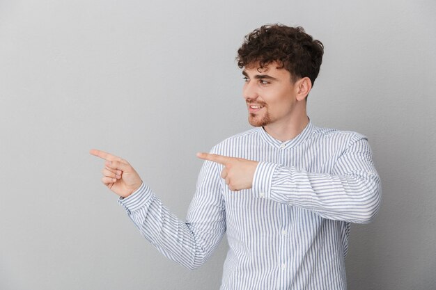 Retrato de hombre rizado optimista vestido con camisa sonriendo y señalando con el dedo a un lado en copyspace aislado sobre pared gris