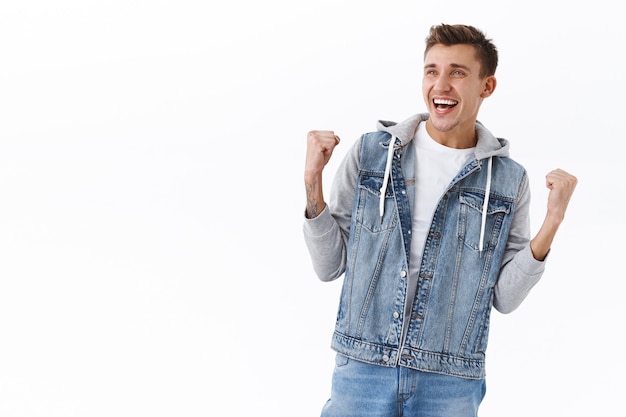 Retrato de hombre rubio alegre y triunfante guapo con chaqueta de mezclilla, bomba de puño lograr el objetivo, mirando la pantalla de televisión en el pub mientras ve la apuesta ganadora de un juego deportivo, convertirse en campeón, celebrando la victoria