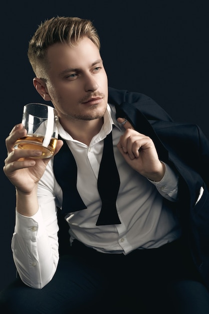 Retrato de hombre rubio guapo en elegante esmoquin y pajarita posando con vaso de whisky en pared negra