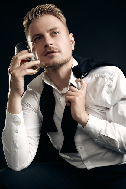 Retrato de hombre rubio guapo en elegante esmoquin y pajarita posando con vaso de whisky en pared negra