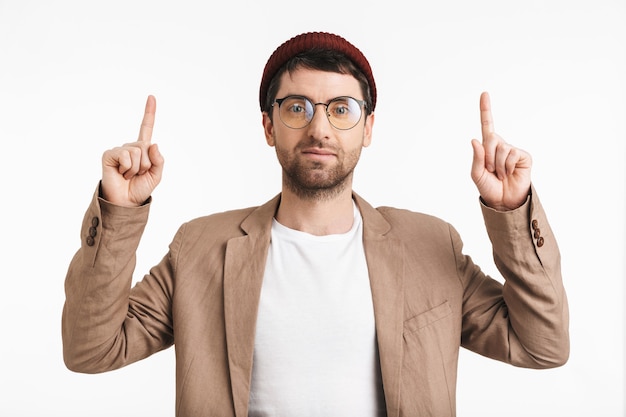 Retrato de hombre seguro con rastrojo con sombrero y anteojos apuntando con el dedo hacia arriba en el copyspace aislado sobre la pared blanca