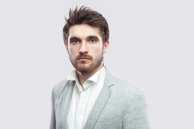 Retrato de hombre serio barbudo guapo con pelos castaños y barba en camisa blanca y traje gris casual de pie y mirando a cámara. tiro del estudio de interior, aislado en fondo gris claro.