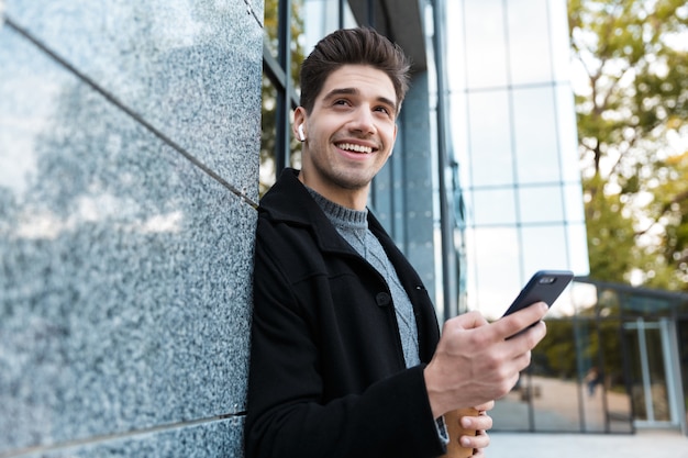 Retrato de hombre sonriente de 30 años con auriculares con teléfono inteligente y café para llevar mientras está de pie frente al edificio de cristal