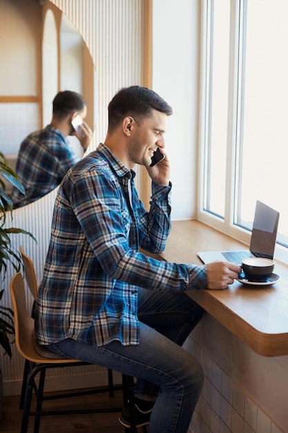 Retrato de un hombre sonriente fandsome de 30 años hablando por teléfono y trabajando de forma remota en una computadora portátil