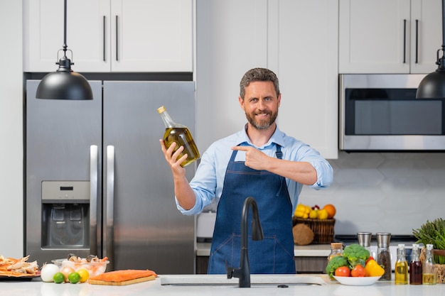 Retrato de hombre sonriente feliz en delantal de cocinero preparando comida natural con botella de aceite de oliva en la cocina