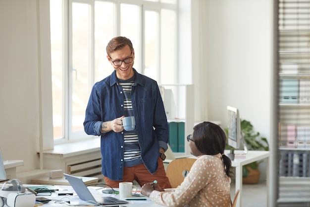 Retrato de hombre sonriente hablando con un colega afroamericano mientras disfruta de una pausa para el café en la moderna oficina blanca, espacio de copia