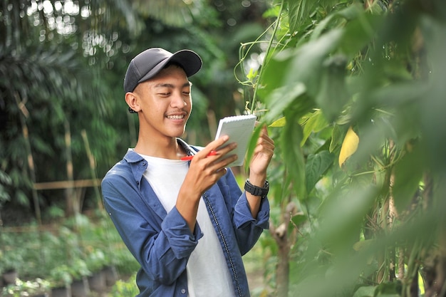 Retrato de hombre sonriente joven agricultor asiático comprobando el pequeño cuaderno. Feliz joven agricultor asiático en el jardín, en el lugar.