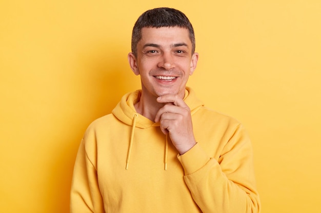 Retrato de un hombre sonriente y satisfecho con una capucha de estilo informal posando aislada sobre fondo amarillo mirando la cámara con expresión positiva sosteniendo la barbilla