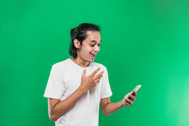 Retrato hombre sosteniendo y mirar telefono con expresión de felicidad sorprendida mientras levanta una mano, aislado sobre fondo verde