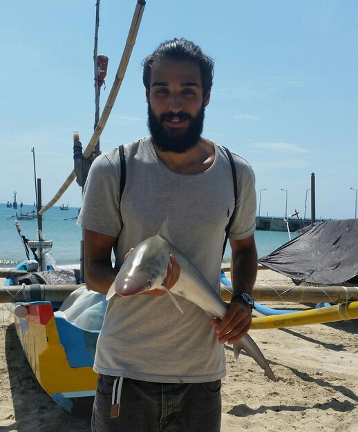 Foto retrato de un hombre sosteniendo un pez mientras está de pie en la playa contra el cielo