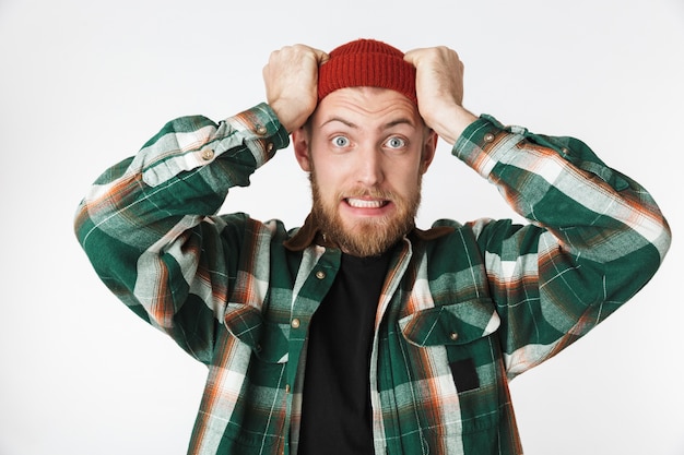 Retrato de hombre tenso con sombrero y camisa a cuadros gritando y agarrando la cabeza, mientras está de pie aislado sobre fondo blanco.