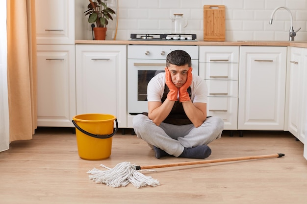 Retrato de un hombre triste y molesto que usa un delantal de ropa informal y guantes de goma naranja sentados en el piso de la cocina con las piernas cruzadas se ve cansado y agotado limpiando el hogar