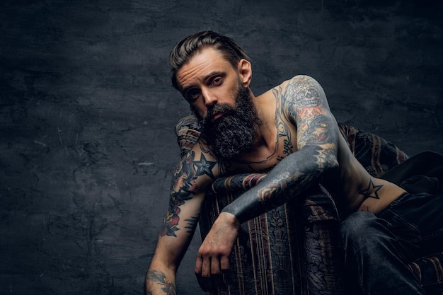 Retrato de un hombre triste tatuado desesperado, que está sentado en un sillón en un escondite oscuro.