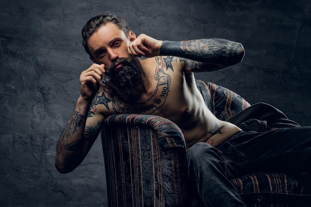 Retrato de un hombre triste tatuado desesperado, que está sentado en un sillón en un escondite oscuro.