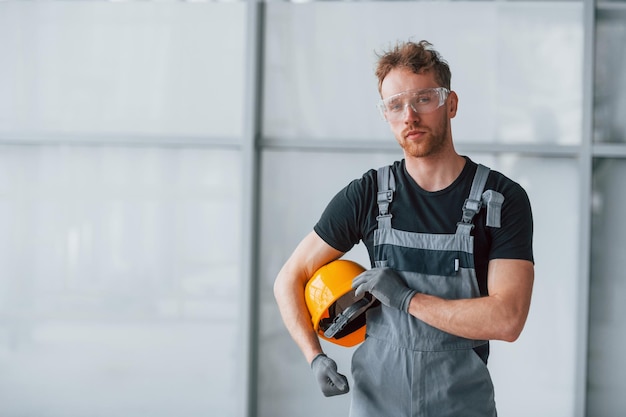 Retrato de un hombre con uniforme gris y un casco naranja en las manos que se encuentra en el interior de una gran oficina moderna durante el día