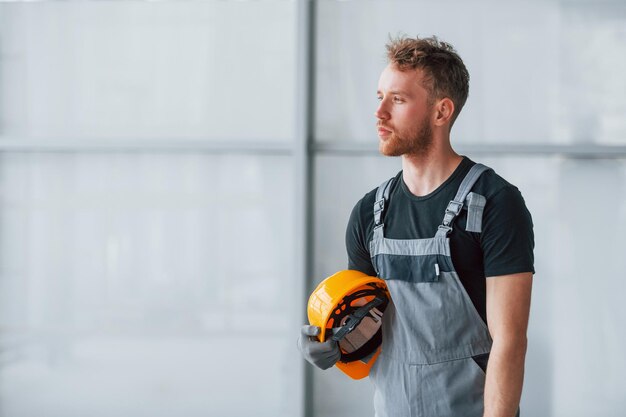 Retrato de un hombre con uniforme gris y un casco naranja en las manos que se encuentra en el interior de una gran oficina moderna durante el día