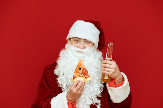 Retrato hombre vestido como Santa Claus sosteniendo una botella de cerveza y una rebanada de pizza