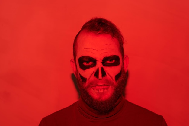Retrato de hombre zombie barbudo enojado con maquillaje sombrío de pie en concepto de halloween de luz roja