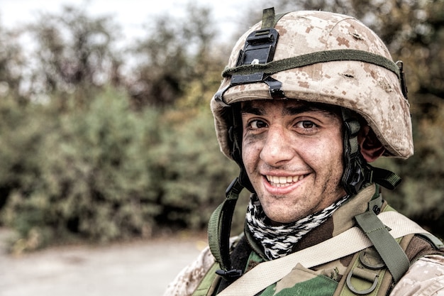 Retrato de hombro de feliz sonriente joven soldado en casco de batalla con arañazos en camuflaje