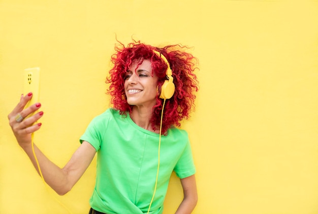 Retrato horizontal de uma mulher latina com cabelo vermelho afro segurando um telefone celular e curtindo ouvir música com fones de ouvido amarelos sobre fundo amarelo isolado