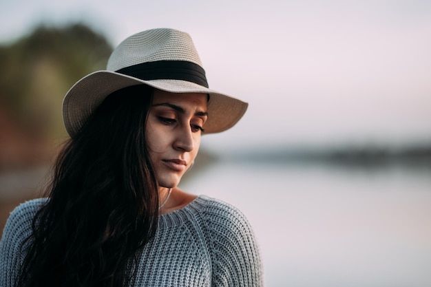 Retrato horizontal de uma mulher pensativa e séria com os olhos fechados usando um chapéu por um lago
