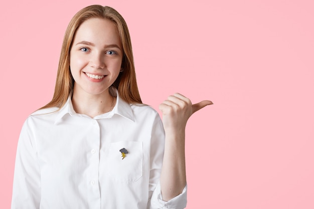 Retrato horizontal de feliz mujer caucásica indica con el pulgar en blanco